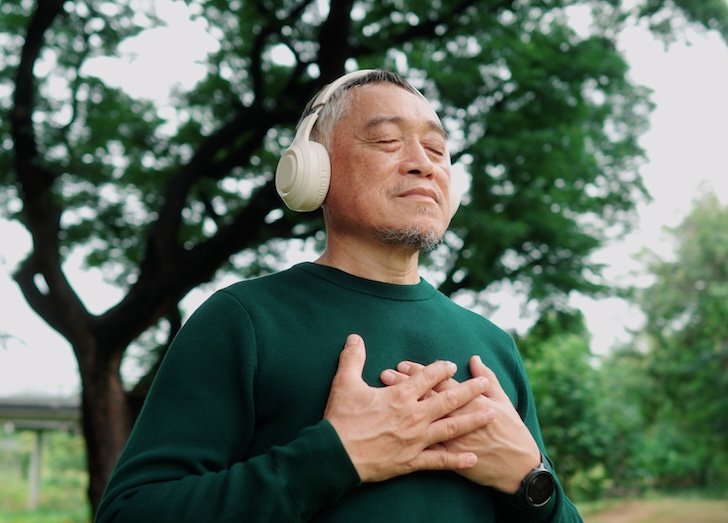 Man meditating under a tree with wireless headphones, calm and refreshed.